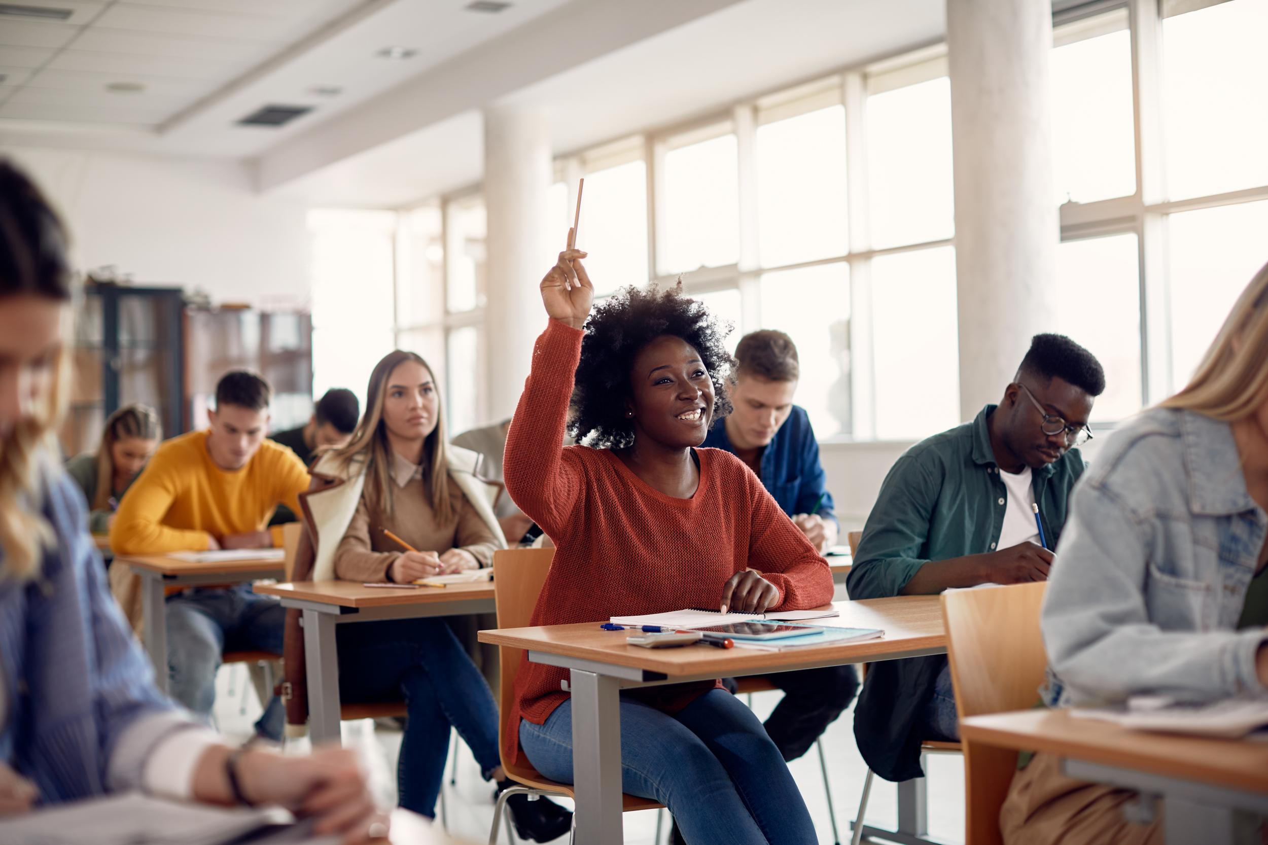 A young woman smiles and raises her hand in a college-age classroom full of students